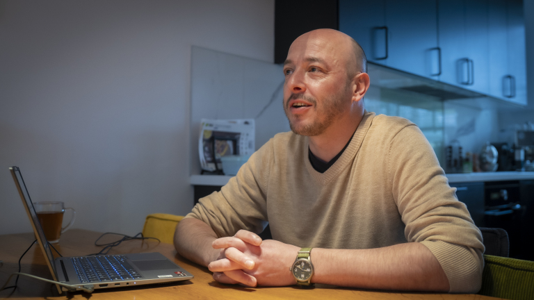 Man aan tafel met een laptop en een kopje thee, kijkend naar links, in een keukenomgeving.