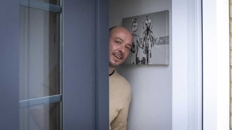 Man gluurt lachend om de hoek van een deur met een zwart-witfoto van kamelenrijders op de achtergrond.