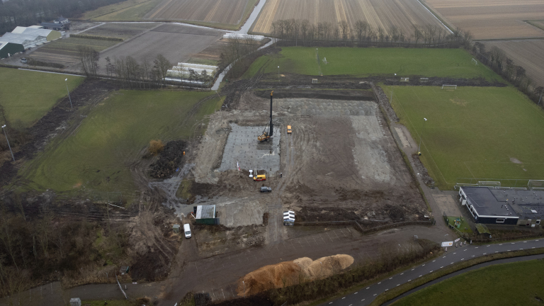 Luchtfoto van een bouwterrein met een boormachine, voertuigen en aangrenzende sportvelden in een landelijk gebied.