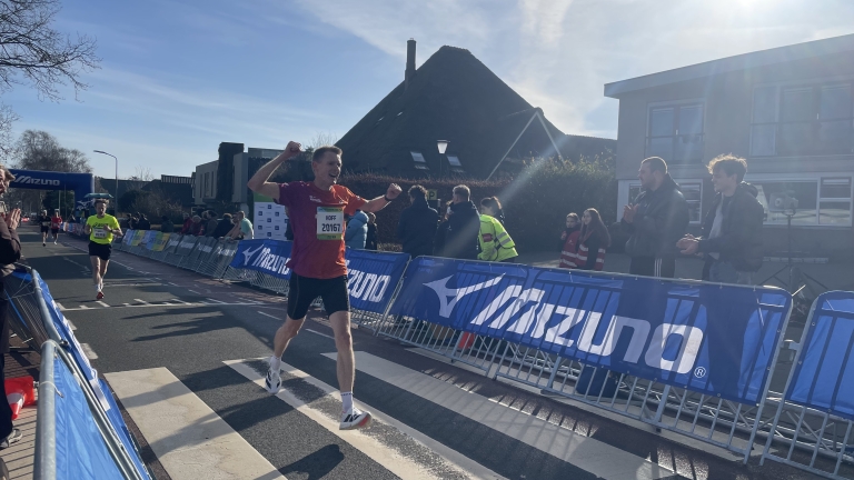 Een hardloper in een rood shirt steekt juichend de finishlijn over, terwijl omstanders klappen langs een met hekken afgebakende baan.