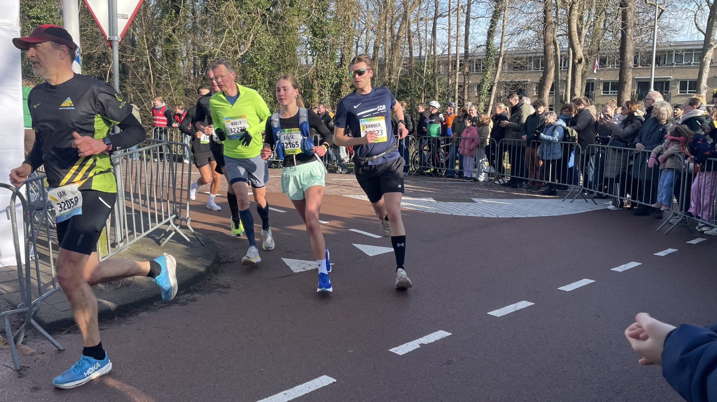 Verschillende hardlopers race langs een parcours, omgeven door toeschouwers achter hekken op een zonnige dag.