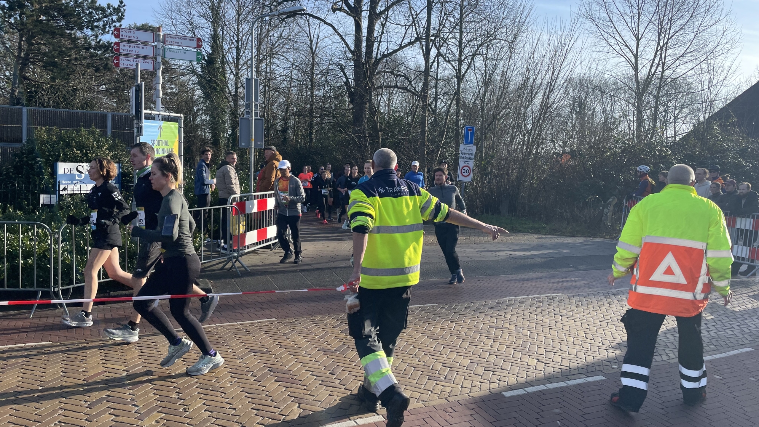 Hardlopers tijdens een wedstrijd op een zonnige dag, begeleid door verkeersbegeleiders in gele en oranje jassen.