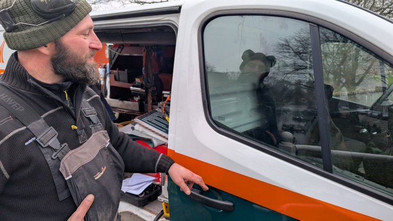 Man in werkkleding opent schuifdeur van een bestelwagen, met een hond op de passagiersstoel.