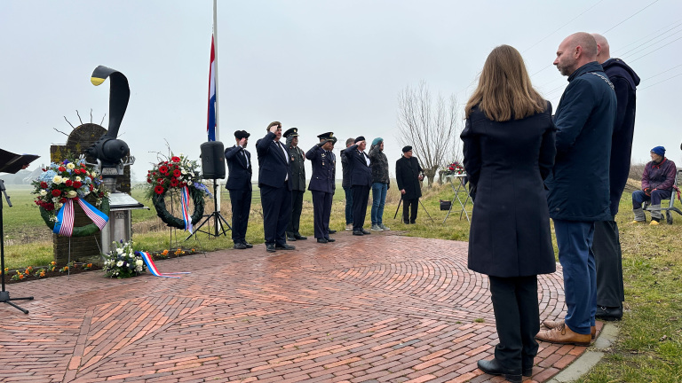 Mensen in uniform wonen een herdenking bij bij een monument, omringd door kransen en vlaggen.