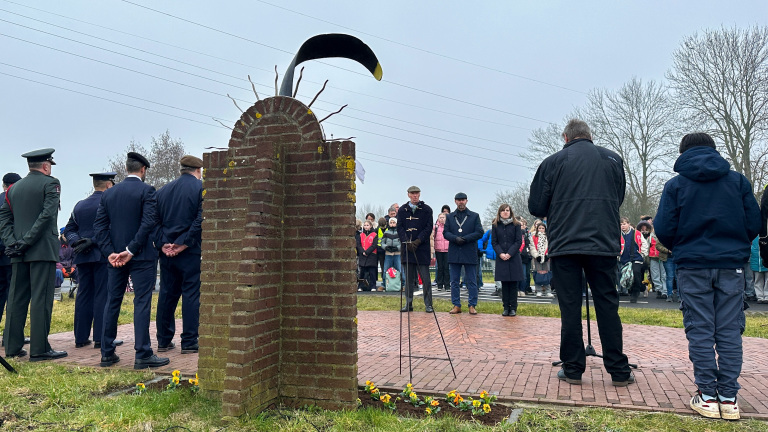 Mensen verzamelen zich voor een ceremonie bij een bakstenen monument met een herdenkingskrans, met bloemen op de grond.