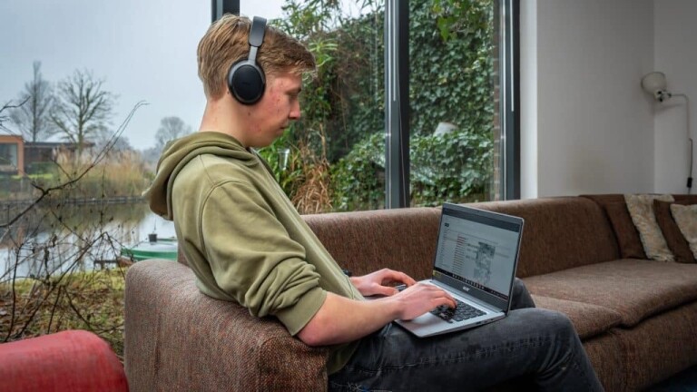 Persoon met koptelefoon werkt op laptop zittend op een bank, met uitzicht op een winterse tuin door het raam.