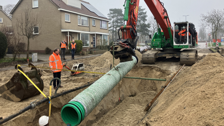 Bouwvakkers installeren een grote groene buis in een greppel met behulp van een graafmachine in een woonwijk.