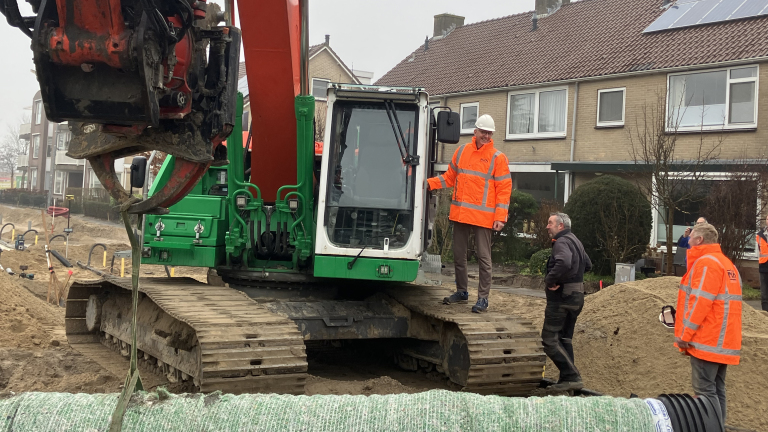 Bouwvakkers in oranje veiligheidskleding staan bij een groene graafmachine op een bouwplaats in een woonwijk.
