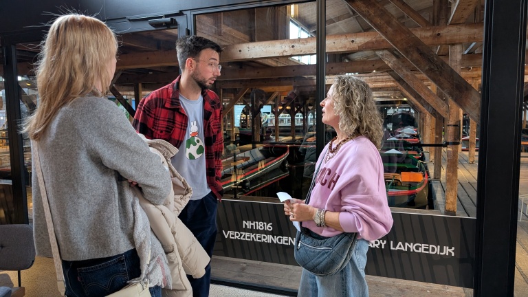 Drie mensen in gesprek in een houten gebouw met boten op de achtergrond.