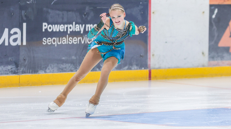 Kunstschaatsster in een blauwe en groene outfit maakt een bocht op het ijs, gefotografeerd tijdens een wedstrijd.