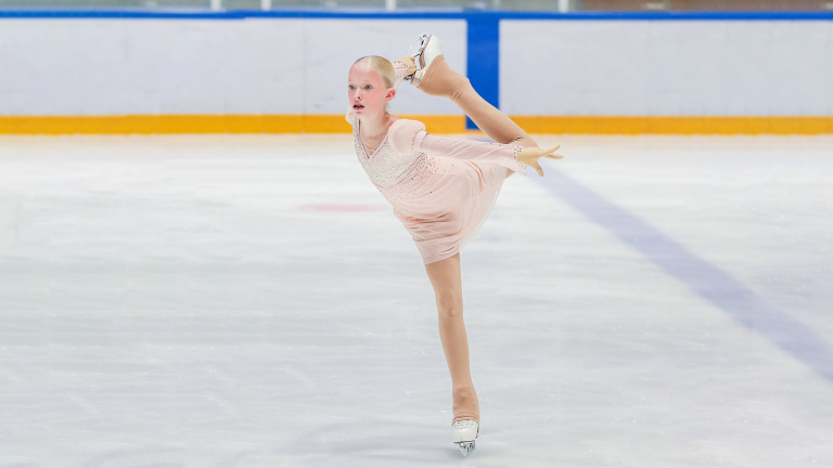 Een jonge kunstschaatsster voert een pirouette uit op de ijsbaan, gekleed in een lichtroze jurk.