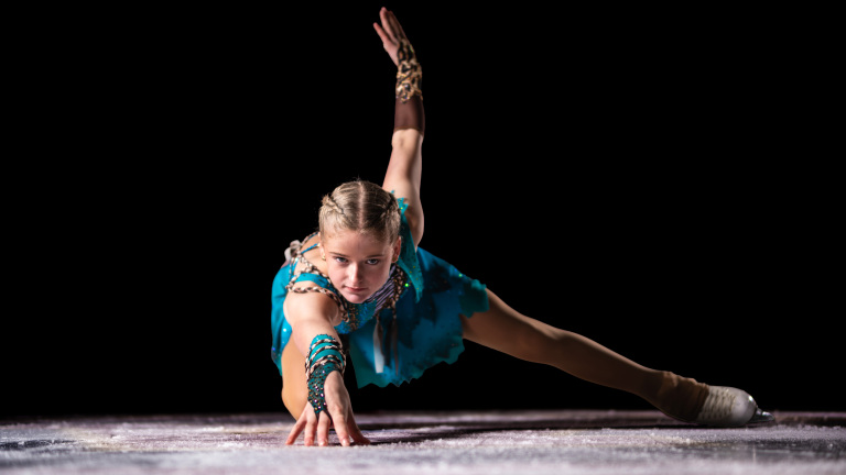 Kunstschaatser in blauwe jurk doet een artistieke pose met een uitgestrekte arm op het ijs.