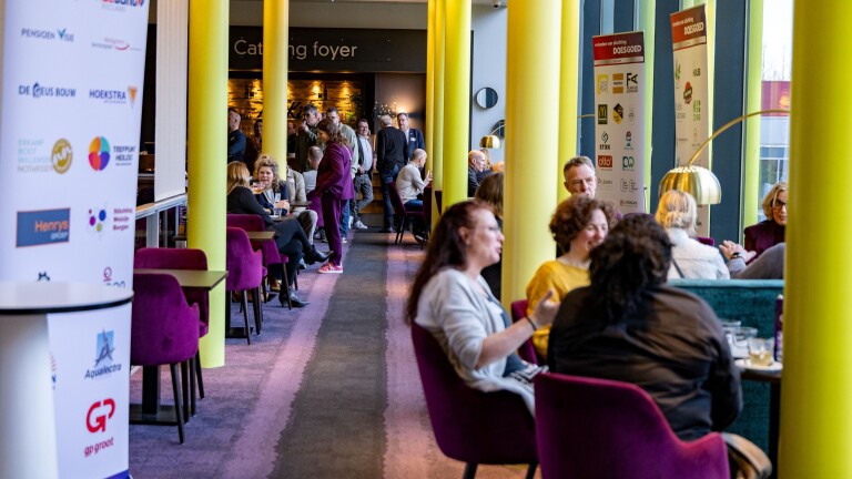 Mensen zitten en staan in een lobby met paarse stoelen en gele pilaren, waar spandoeken met bedrijfslogo's te zien zijn.