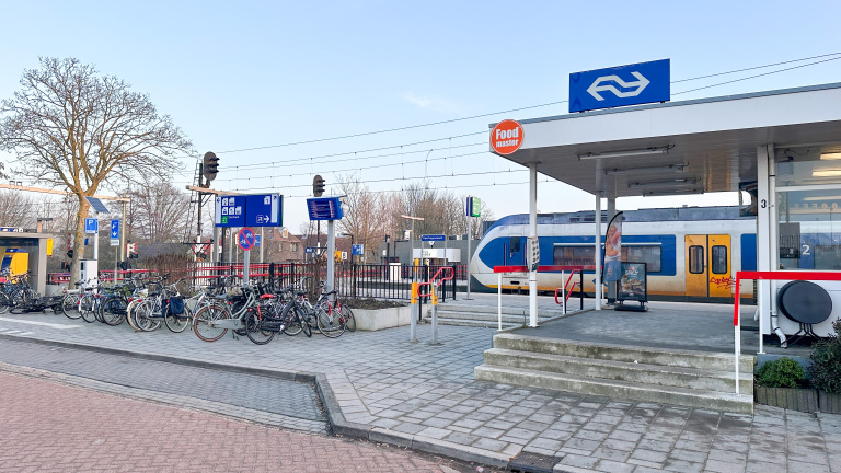 Een treinstation met NS-logo, fietsenstalling, en een trein op het perron; borden geven richtingen aan.