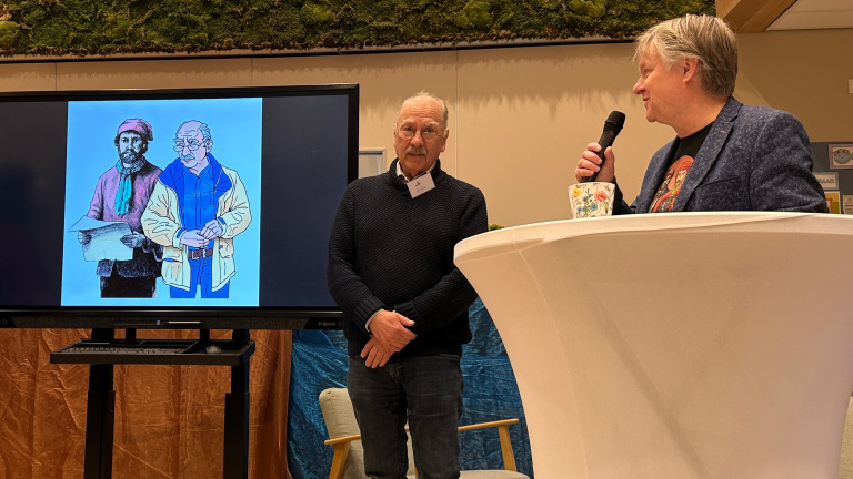 Twee mannen staan naast een spreker met een microfoon bij een tafel met een getekende afbeelding op een scherm.