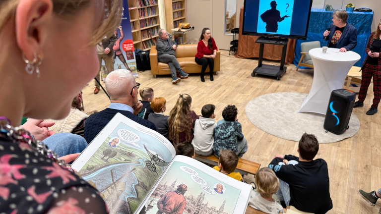 Een meisje leest een stripboek terwijl een presentatie met een projectiescherm plaatsvindt in een bibliotheek.