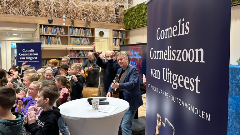 Kinderen en volwassenen in een bibliotheek applaudisseren tijdens een muzikaal optreden, met banners over Cornelis Corneliszoon van Uitgeest.
