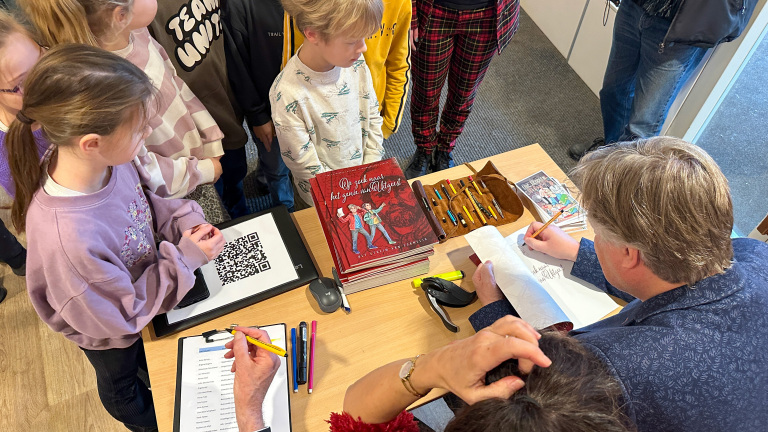 Een auteur signeert een kinderboek omringd door een groep kinderen, met een stapel boeken en schrijfgerei op tafel.