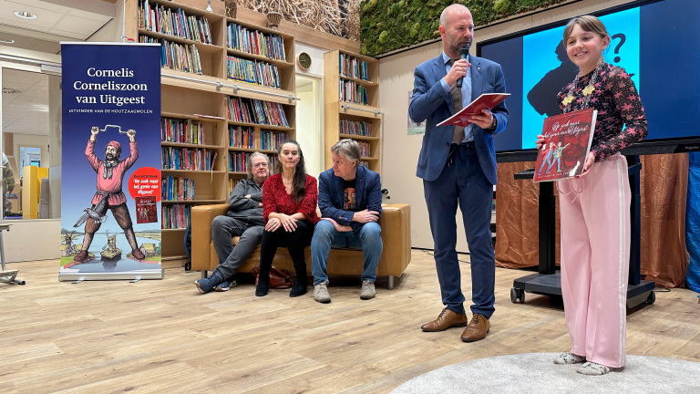 Een presentator en een meisje met een boek staan voor een groep mensen, naast een roll-up over Cornelis Cornelisz. van Uitgeest.