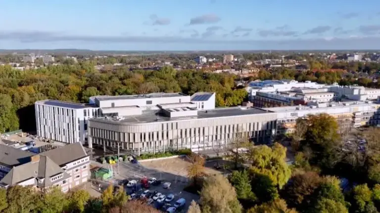 Groot, modern gebouw omringd door bomen, met een parkeerplaats aan de voorkant onder een heldere, blauwe lucht.