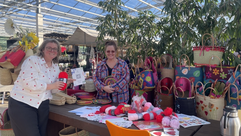 Twee vrouwen glimlachen achter een tafel vol kleurrijke manden, garen en decoraties in een kas met planten.