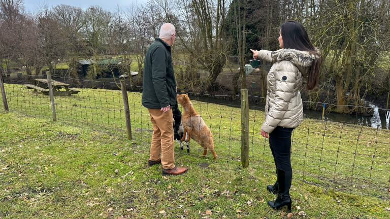 Twee mensen en twee geiten staan bij een hek, terwijl een vrouw met jas ergens naartoe wijst in een landelijke omgeving.