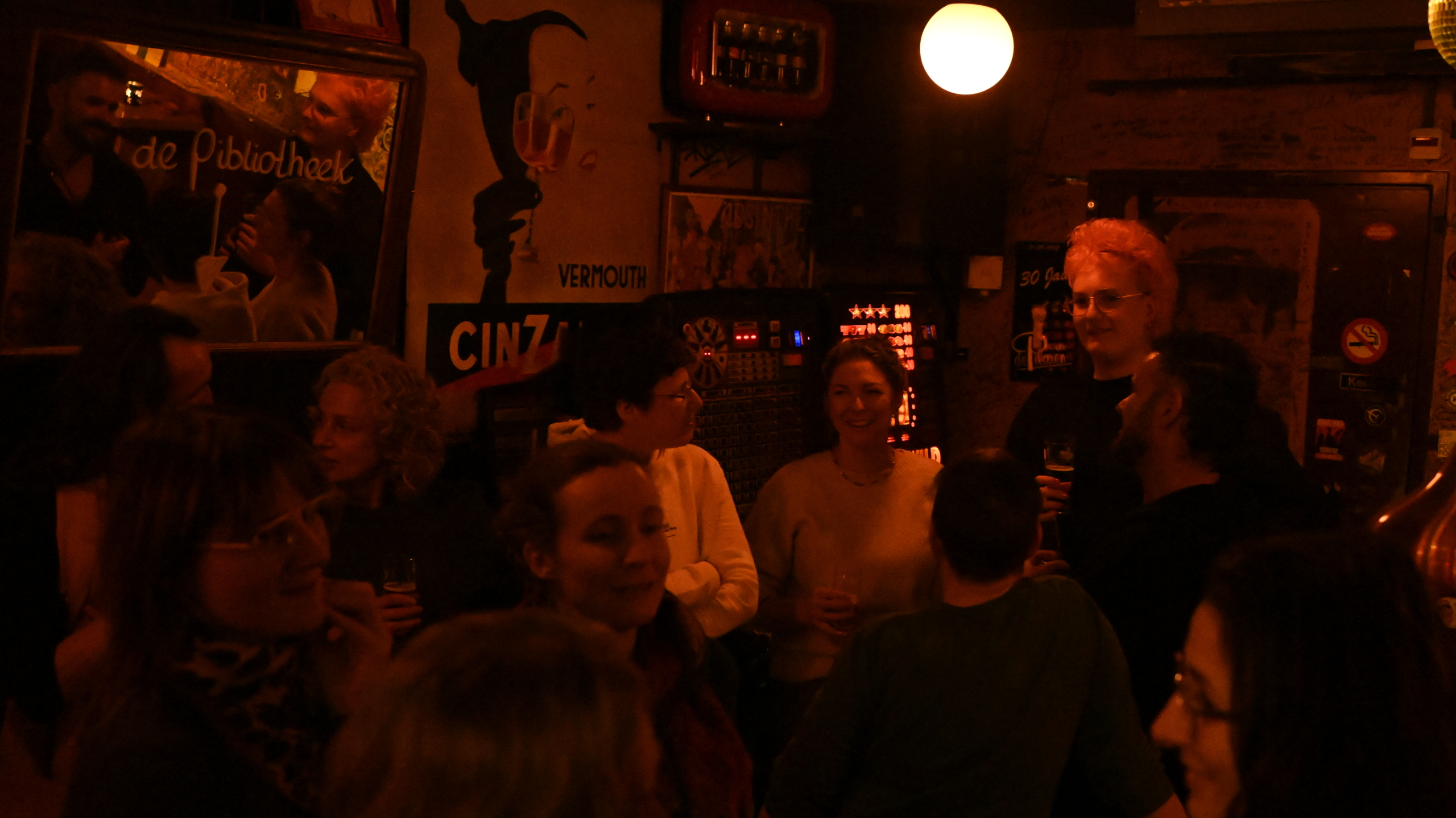 Een groep mensen staat in een sfeervolle bar met warme, gedimde verlichting en posters aan de muur.