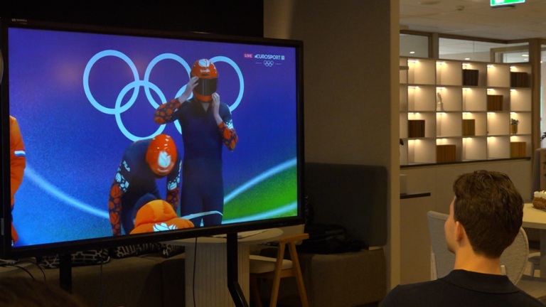 Persoon kijkt naar een tv-scherm dat een Olympische speedskatewedstrijd uitzendt, in een ruimte met een taart op tafel.