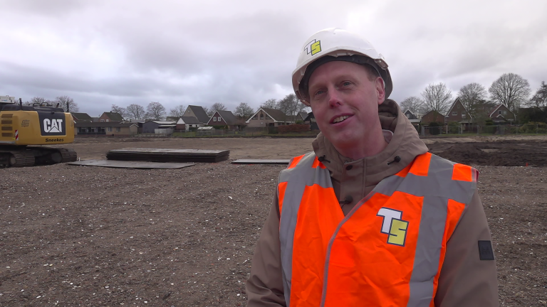 Man met witte bouwhelm en fluorescerend oranje vest op een bouwterrein, met huizen en een graafmachine op de achtergrond.