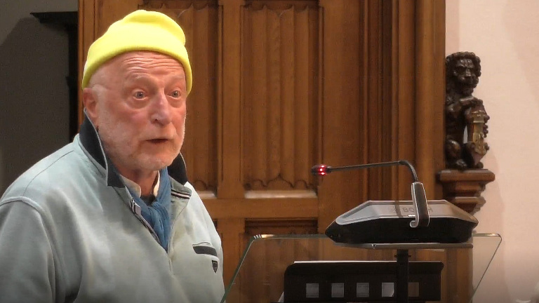 Man met een gele muts staat bij een spreekgestoelte met een microfoon, in een houten interieur.