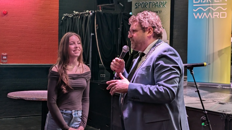 Een man in een pak spreekt met een microfoon tegen een jonge vrouw die glimlacht, tegen de achtergrond van banners.