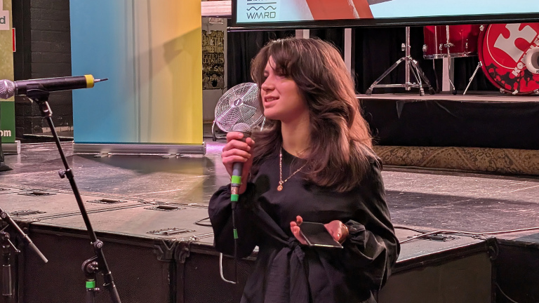 Een vrouw spreekt in een microfoon op een podium, met muziekinstrumenten en schermen op de achtergrond.