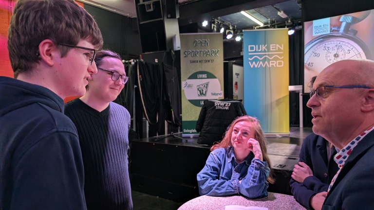 Vier mensen zijn in gesprek rondom een tafel in een zaal met banners van "Open Sportpark" en "Dijk en Waard" op de achtergrond.