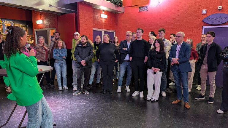 Een vrouw in een groene blazer spreekt voor een groep mensen in een zaal met rode muren.