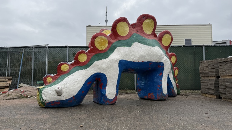 Kleurrijk beeld van een dinosaurus met rode, gele en groene details, staat op een betonoppervlak bij een hek.