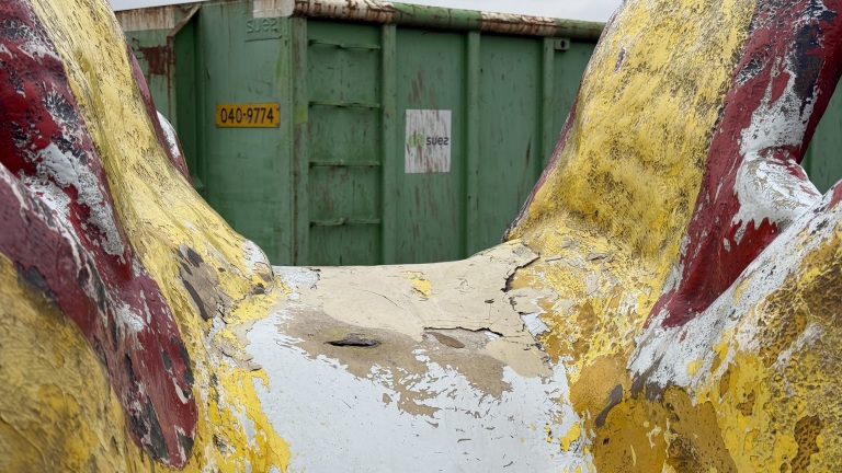 Een verweerd geel en rood gekleurd object met afgebladderde verf, voor een groene container met het label 'suez'.