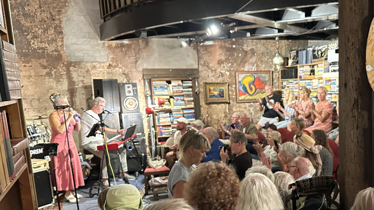 Muzikanten spelen in een gezellige ruimte vol boeken en kunst, omringd door een applauderend publiek.