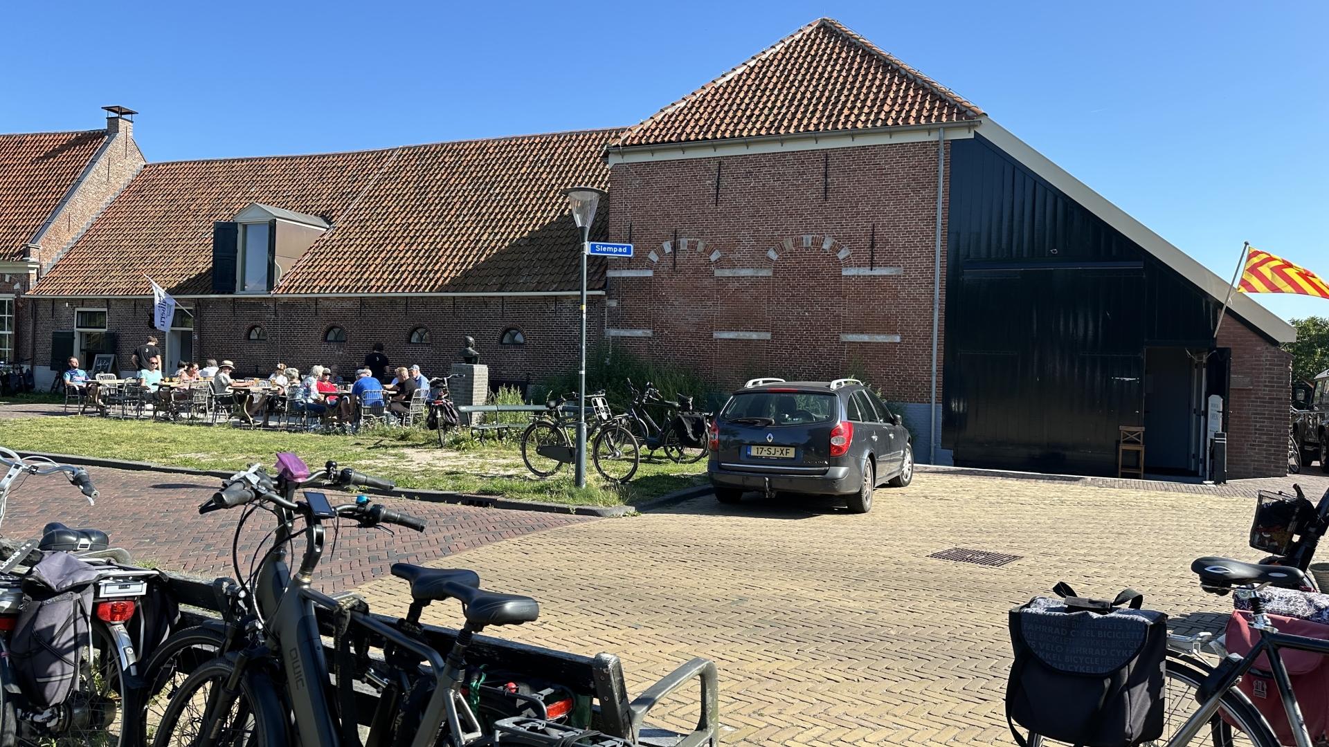 Bakstenen gebouw met terras waar mensen buiten zitten, omringd door geparkeerde fietsen en auto's, en een vlag.