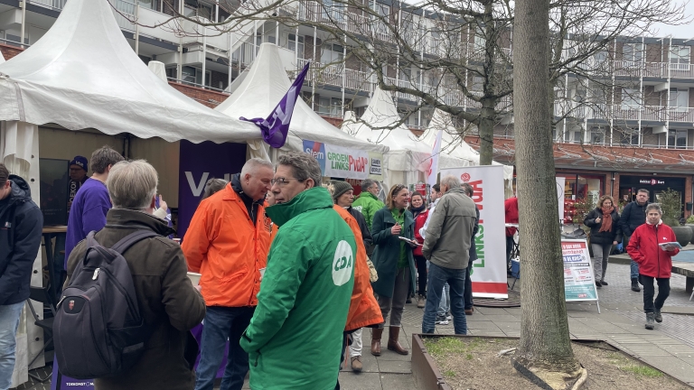 Mensen bij politieke campagnekraampjes met banners van verschillende partijen op een plein.