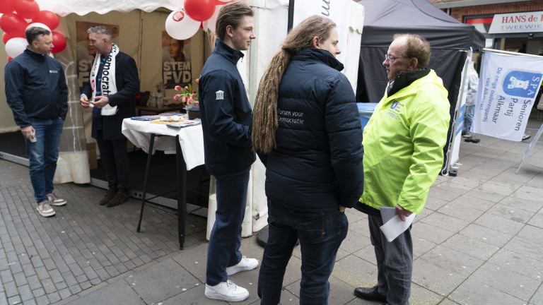 Mensen in gesprek bij een politieke stand van Forum voor Democratie met flyers en posters, omringd door ballonnen.