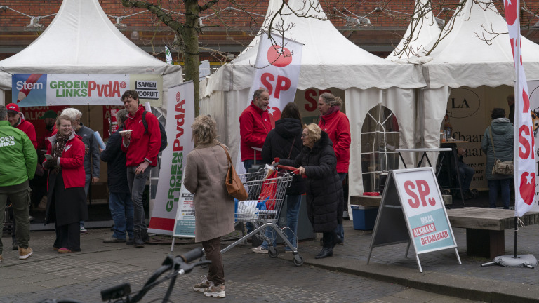 Mensen staan bij promotiestands van politieke partijen GroenLinks, PvdA en SP op een buitenmarkt.