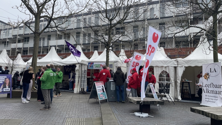 Marktkraampjes met politieke partijen, zoals SP en GroenLinks, en mensen die folders uitdelen op een plein.