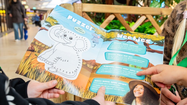 Kinderen lezen een kleurrijk boek in een winkelcentrum met puzzel en weetjes over dieren zoals olifanten en giraffen.