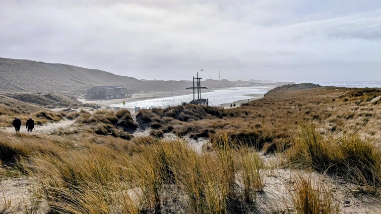 Strand met duinen, een houten gebouw aan de kustlijn en twee wandelende mensen in een grasachtige omgeving.