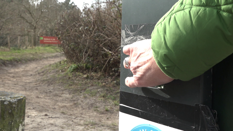 Een hand in een groene jas opent een groene kast naast een onverharde weg met een wazig bord op de achtergrond.