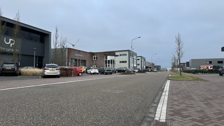 Industriegebied met bedrijfspanden en geparkeerde auto's langs een weg, onder een bewolkte hemel.