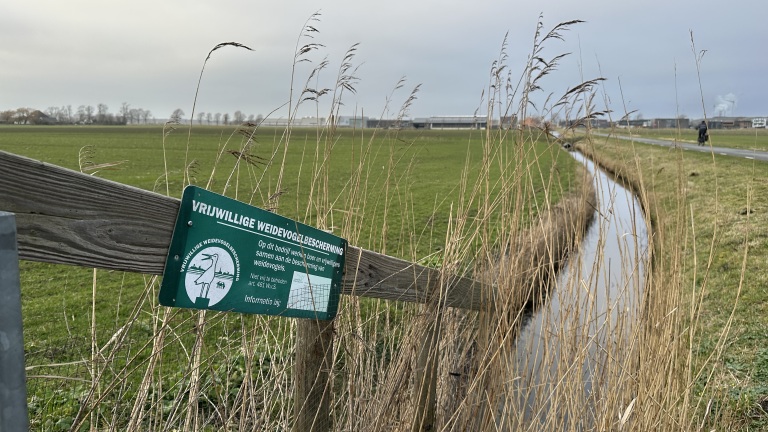 Bordje met "Vrijwillige Weidevogelbescherming" bij een groen weiland met sloot en fietser op de achtergrond.