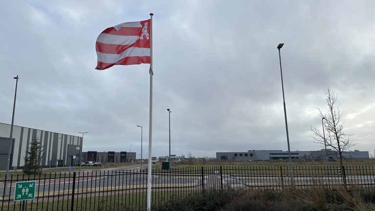 Een rood-wit gestreepte vlag wappert in de wind voor industriële gebouwen onder een bewolkte hemel.