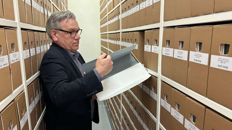 Een man zoekt in een archief een map met documenten tussen rijen van archiefdozen.