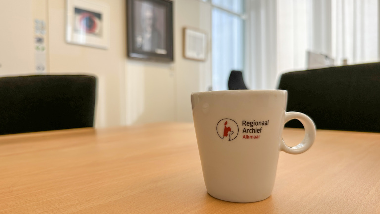 Beker met logo van Regionaal Archief Alkmaar staat op een houten tafel in een kantoorruimte met schilderijen aan de muur.
